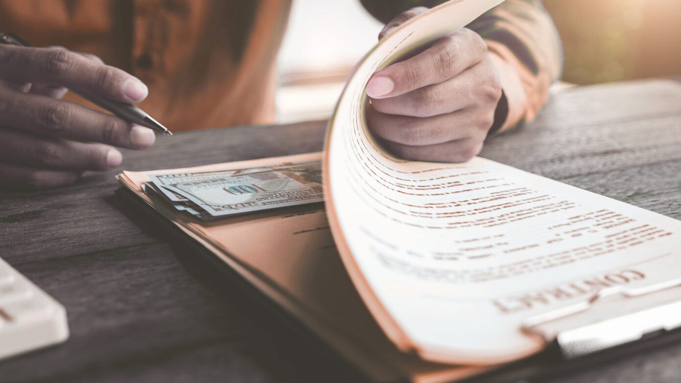 A man in an orange shirt signs a contract on a clipboard which conceals a stack of £100 notes.