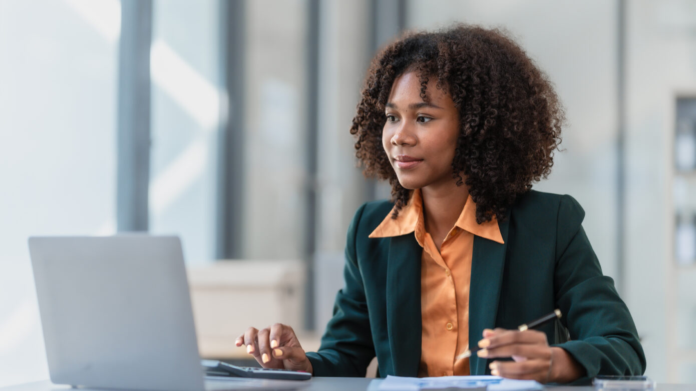 Compliance and governance business woman in an orange shirt and black blazer works on her laptop while holding a pen. There is a printed document on the table in front of her.